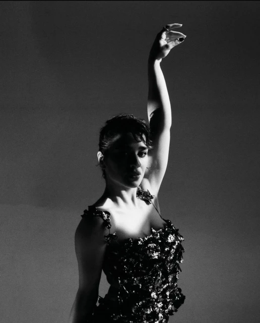 Monochrome portrait of a woman in a textured dress raising her arm in a dramatic pose against a gradient grey backdrop.