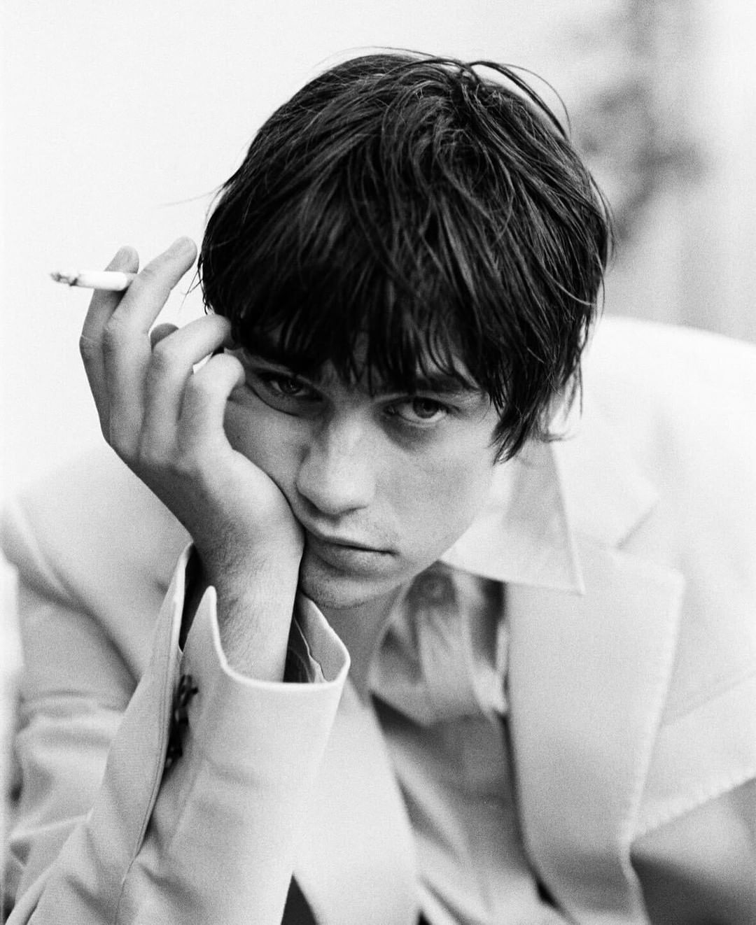Black and white portrait of a young adult male with dark hair holding a cigarette, wearing a suit jacket and button-down shirt.