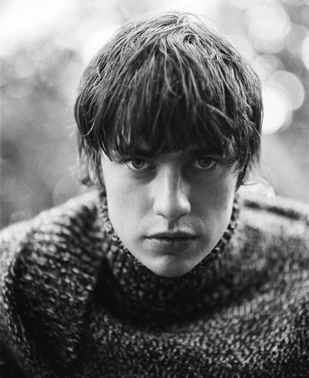 Close-up portrait of a young adult with medium-length brown hair, wearing a knit turtleneck sweater, against a blurred bokeh background.