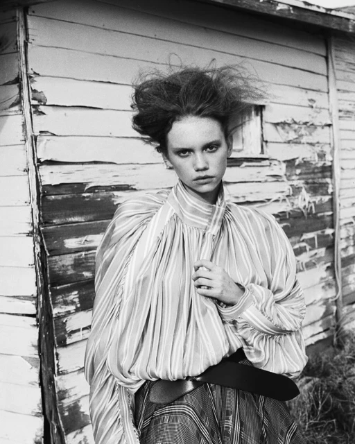 Black and white portrait of a woman with voluminous hair wearing a striped blouse, standing against a weathered wooden wall outdoors.