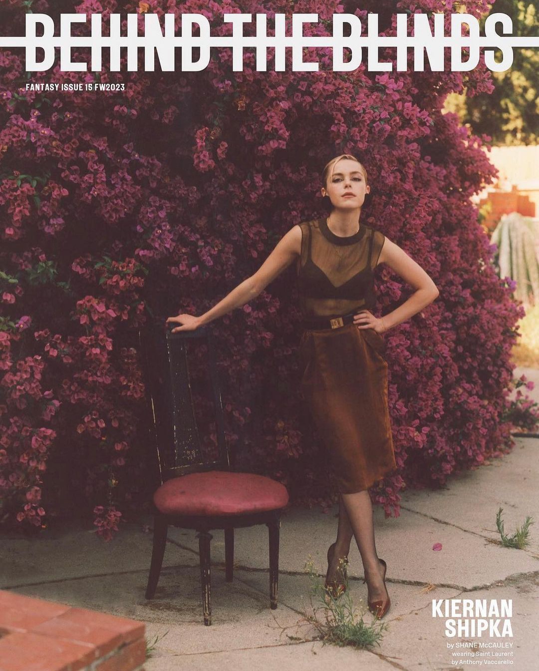 Kiernan Shipka poses on a patio in front of a flowering bush in a fashion editorial for Behind the Blinds Fantasy Issue FW2023.