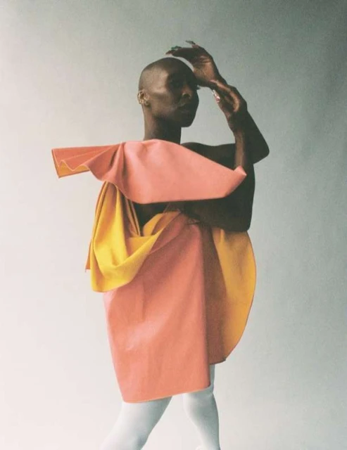 Person in artful orange and yellow garment posing in studio with hands near head; white leggings, neutral studio background, fashion-editorial.