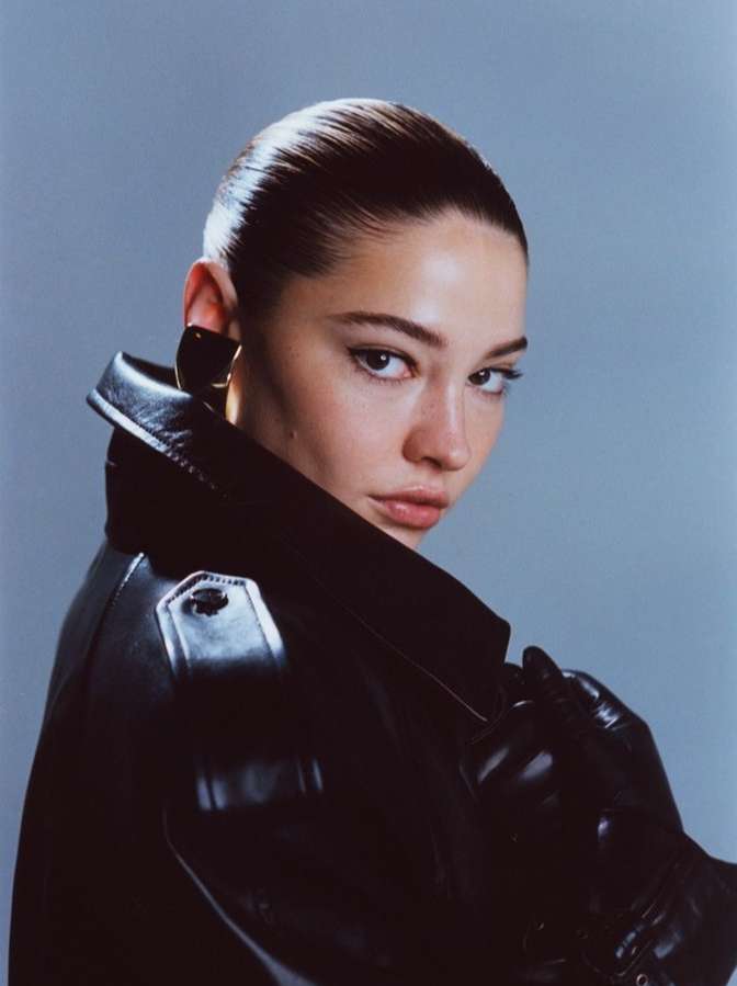 Close-up portrait of a woman with slicked-back hair wearing a black leather coat and gloves against a light blue background.