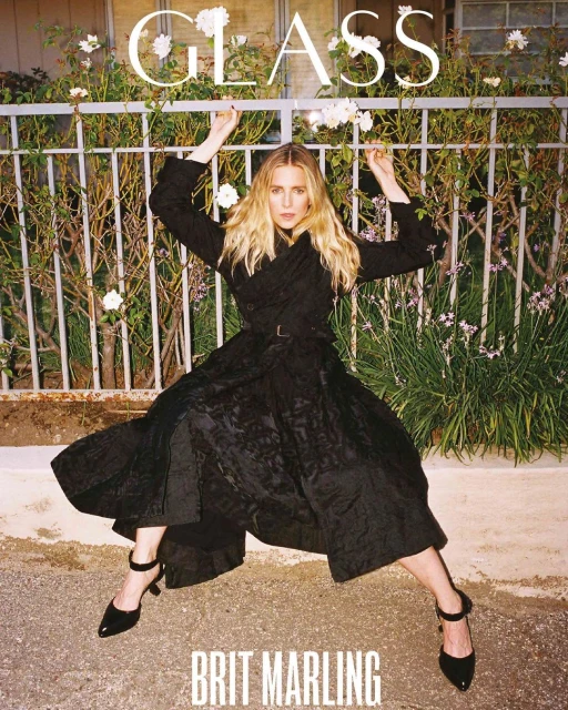 Brit Marling poses outdoors in a black dress and heels against a white fence with climbing flowers for a magazine cover.