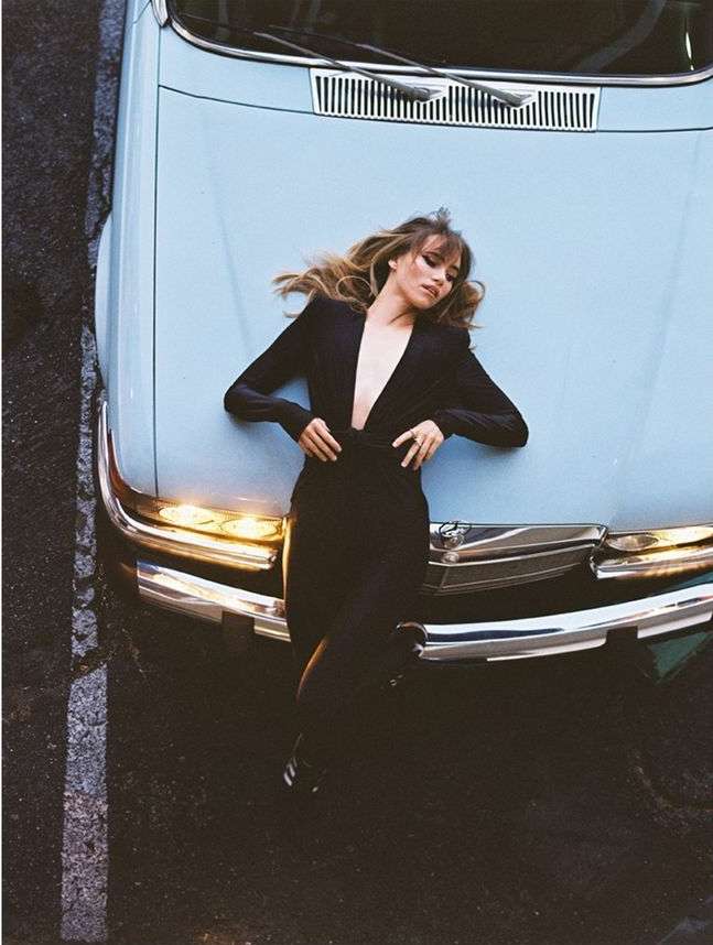 A fashionable adult woman lying on the hood of a vintage light blue car parked on an asphalt street; medium shot from an overhead angle.