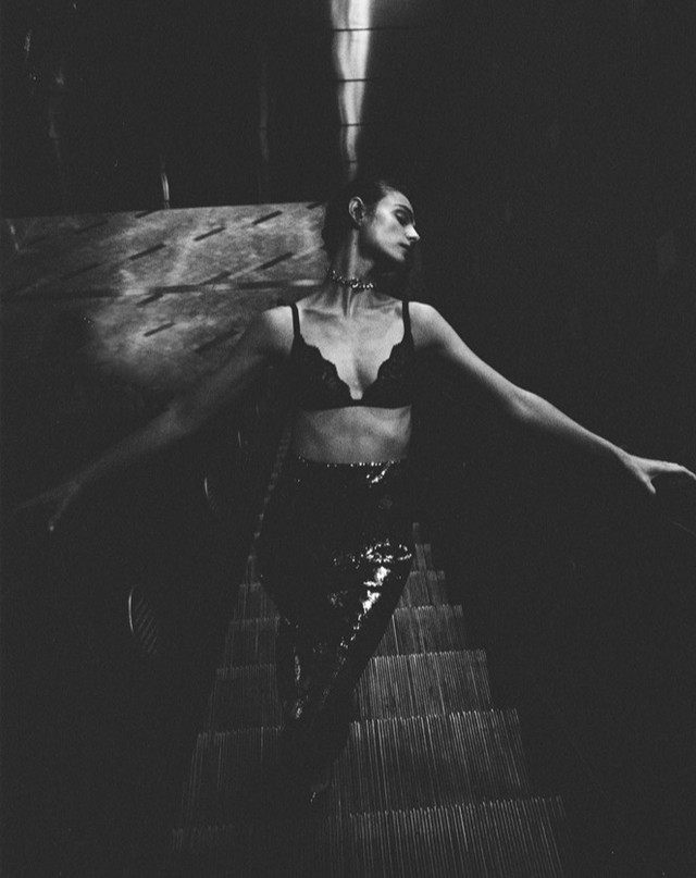 Black and white portrait of a woman in a lace bra and shiny pants posing with outstretched arms on a staircase in a dark setting.