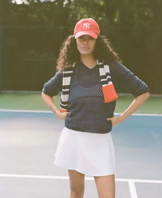 Woman in tennis outfit on court: navy sweater, white skirt, red baseball cap. Posing with hands on hips, tennis court background.