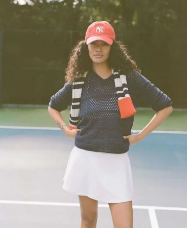 Woman in tennis outfit on court: navy sweater, white skirt, red baseball cap. Posing with hands on hips, tennis court background.