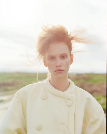 Portrait of a woman with windswept red hair wearing a light cream buttoned coat against an overcast sky and muted landscape.