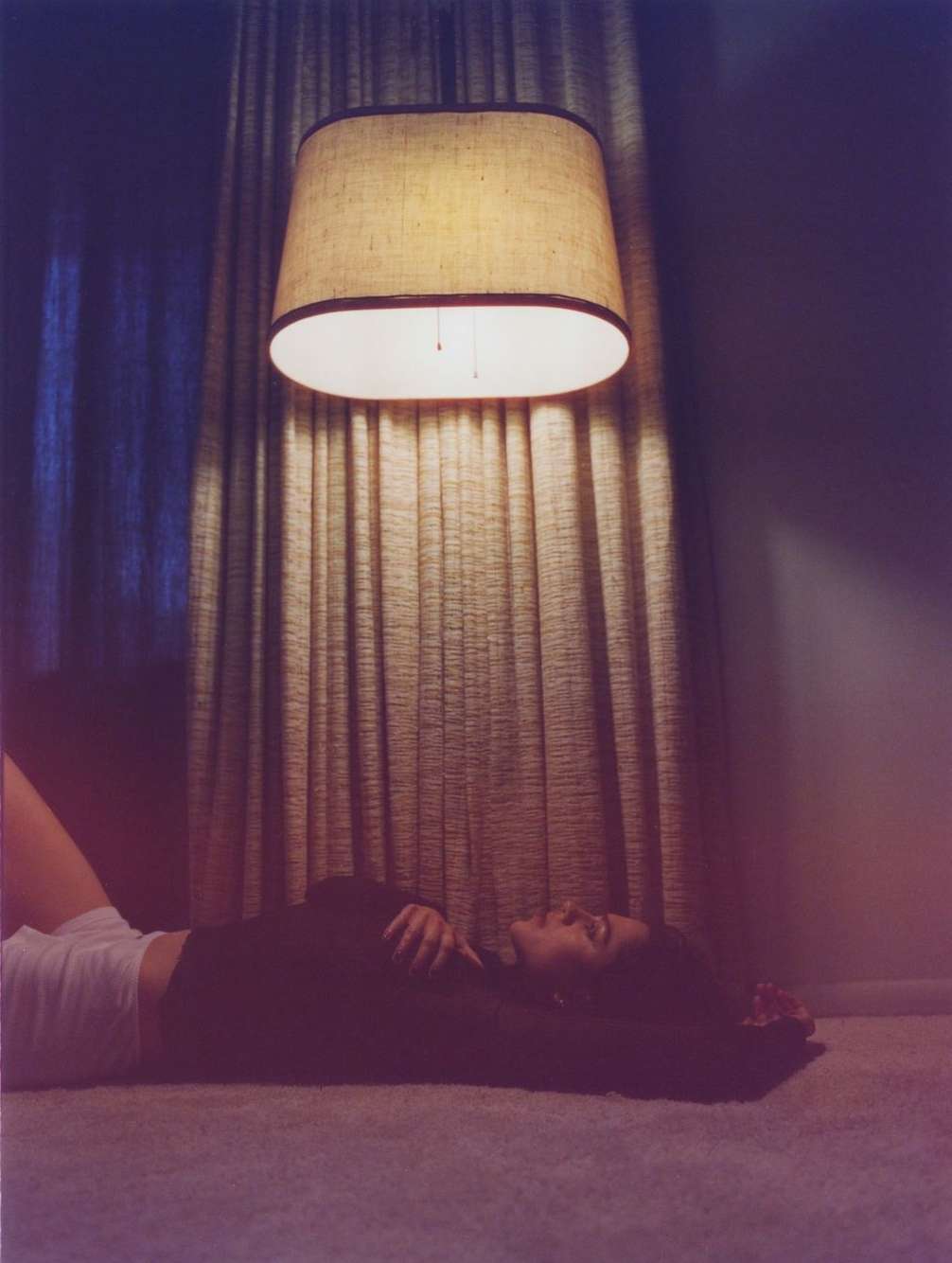 Woman lying on carpet under a lit lamp with a tan shade and beige curtains in the background, shot in an indoor setting.