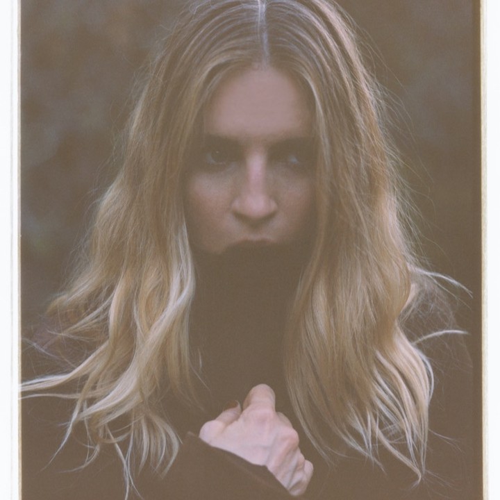 Portrait of a woman with long blond hair, wearing dark clothing, outdoors with a blurred background. The photograph has a washed-out color grade.
