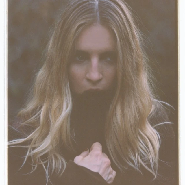 Portrait of a woman with long blond hair, wearing dark clothing, outdoors with a blurred background. The photograph has a washed-out color grade.