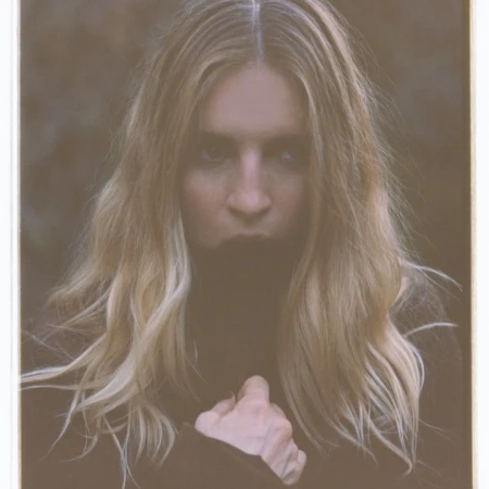 Portrait of a woman with long blond hair, wearing dark clothing, outdoors with a blurred background. The photograph has a washed-out color grade.