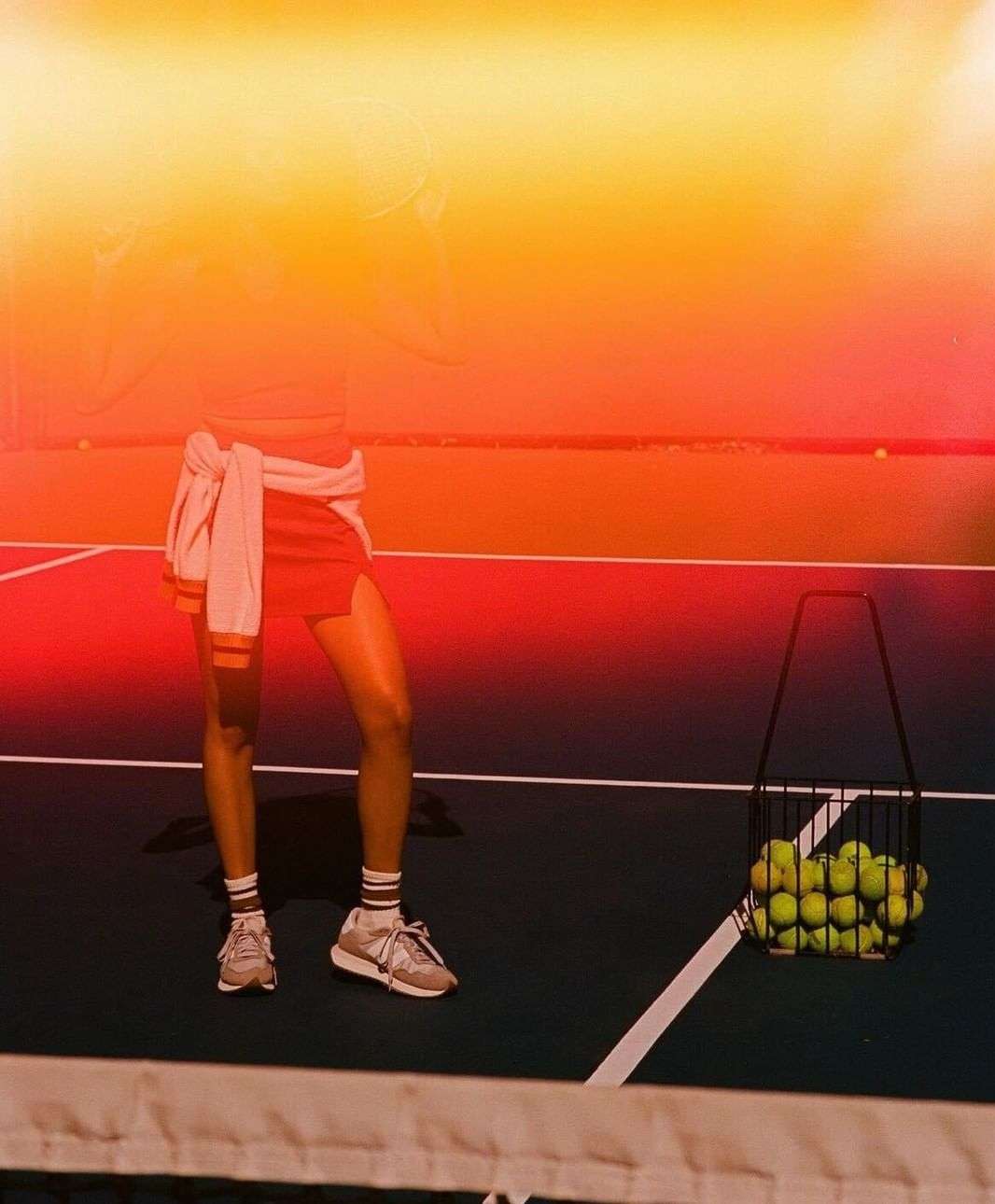 Woman standing on a tennis court near a basket of tennis balls wearing a pink skirt, crop top and sneakers, in bright sunlight.