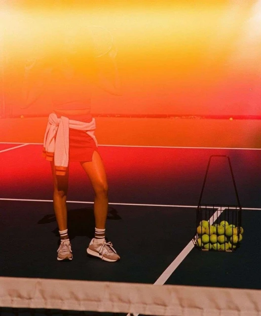 Woman standing on a tennis court near a basket of tennis balls wearing a pink skirt, crop top and sneakers, in bright sunlight.