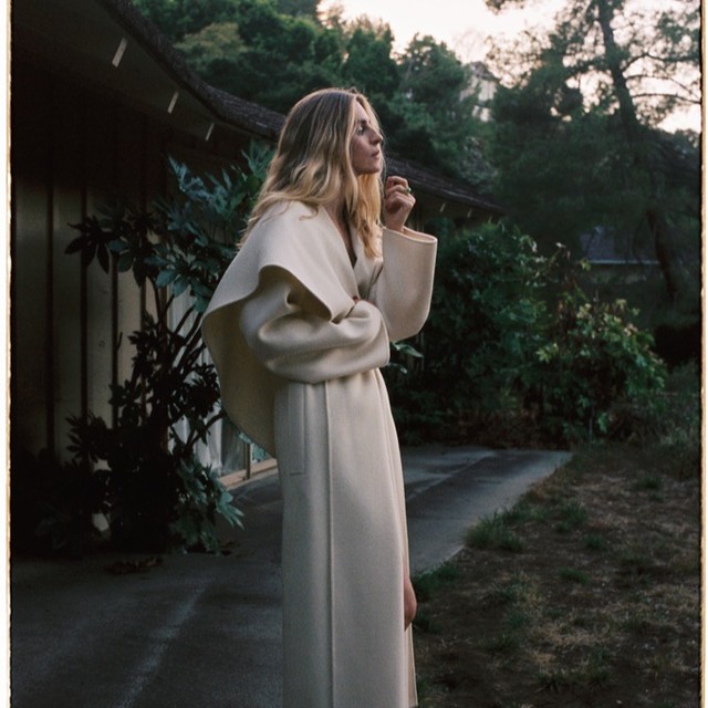 Woman with long blonde hair wearing a long cream coat standing outside a residential building at dusk