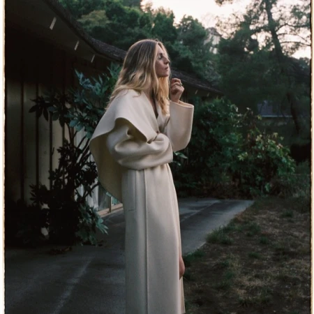 Woman with long blonde hair wearing a long cream coat standing outside a residential building at dusk
