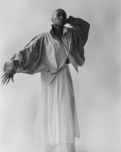 Black and white portrait of an androgynous fashion model posing in an oversized shirt and sheer skirt against a neutral backdrop in a studio.