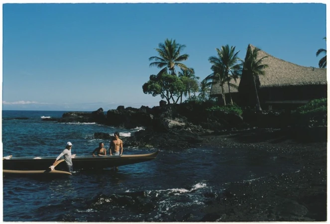 Three people in an outrigger canoe approach a black sand beach bordered by lava rocks, palm trees and a thatched roof building.