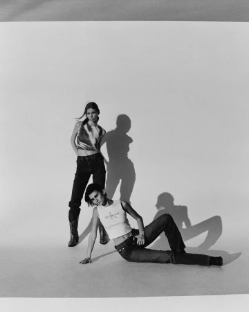Two women pose in a photo studio. One stands wearing a metallic top and jeans, while the other reclines wearing a tank top and jeans.