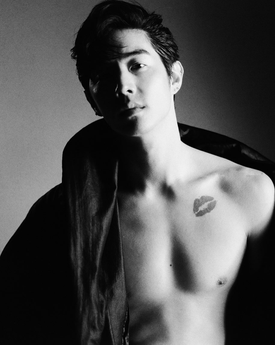 Monochrome portrait of a shirtless adult male with a lip print tattoo, jacket draped on shoulder, against a blurred grey background.