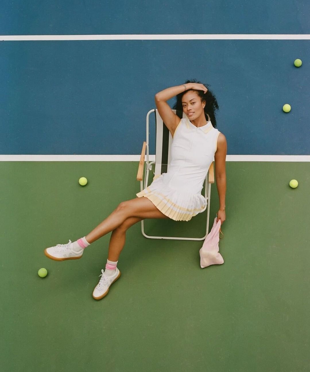 Woman in a tennis dress sits on a folding chair on a blue and green tennis court surrounded by scattered yellow tennis balls overhead shot.