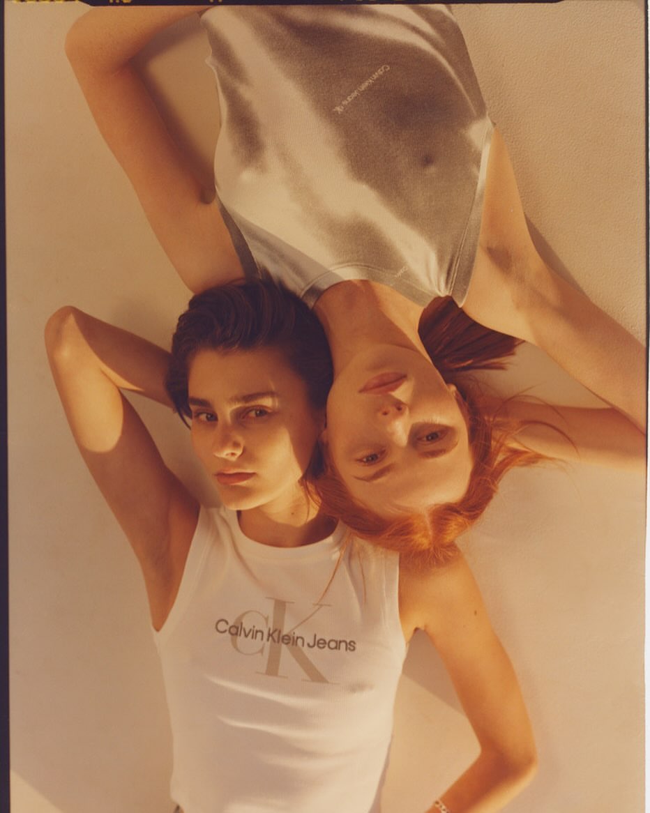 Two women lying down with heads together wearing white Calvin Klein tank tops under warm lighting
