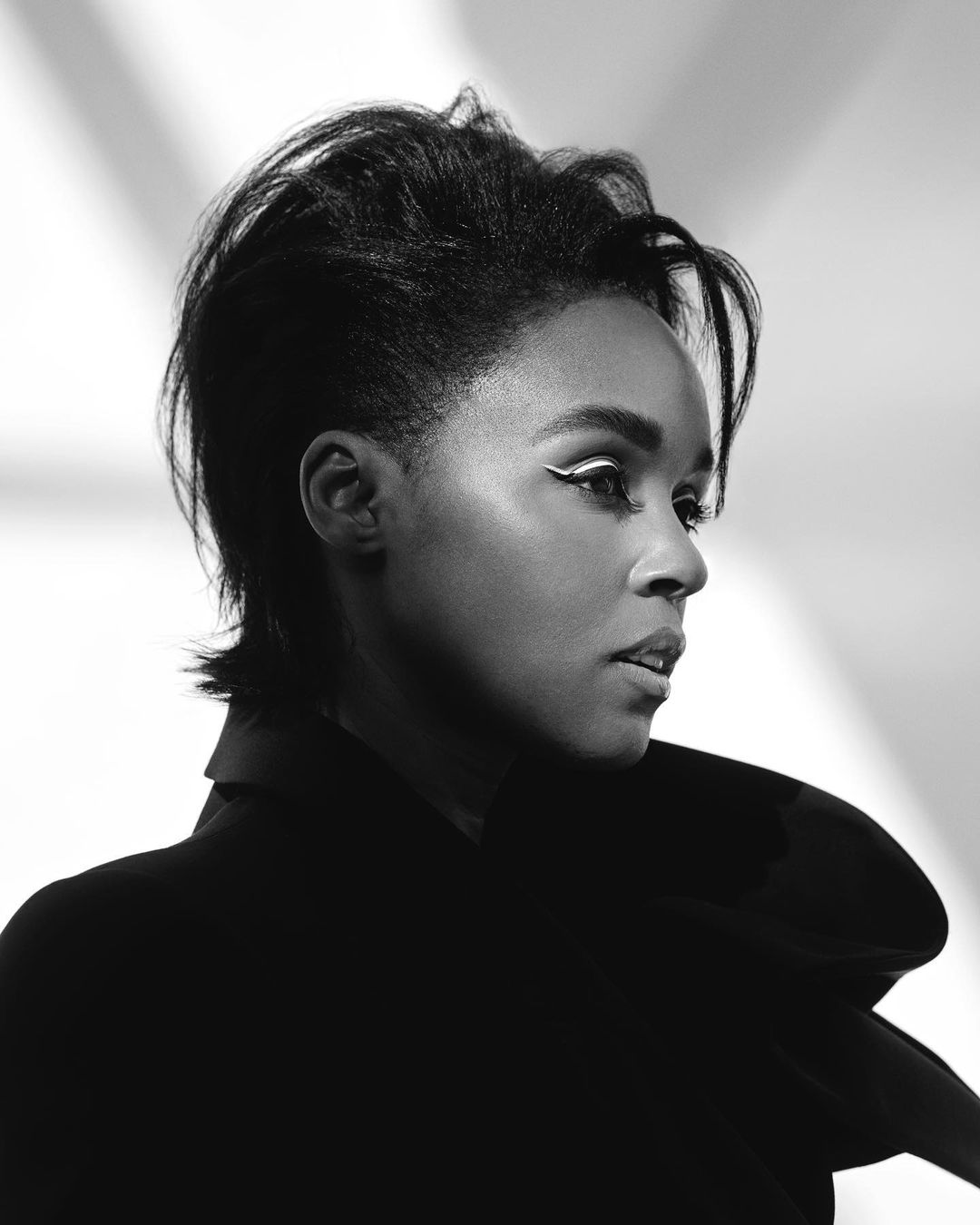Monochrome portrait of a woman with short dark hair, wearing a black blazer and white eyeliner, against a softly lit white backdrop.