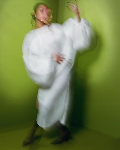 Blurred motion portrait of a female figure in a flowing white dress in a green corner with motion blur emphasizing movement and light.