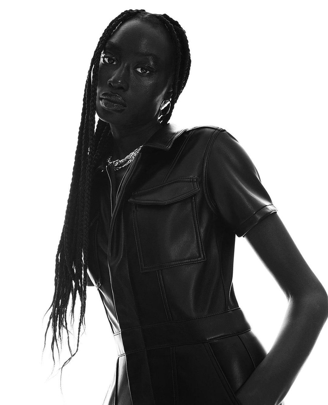 Portrait of a Black woman with long braids wearing a black leather jumpsuit, posing against a bright white background in a fashion shot.
