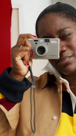 Black woman holding a silver point and shoot compact camera to her eye, wearing a camel and yellow collared shirt.