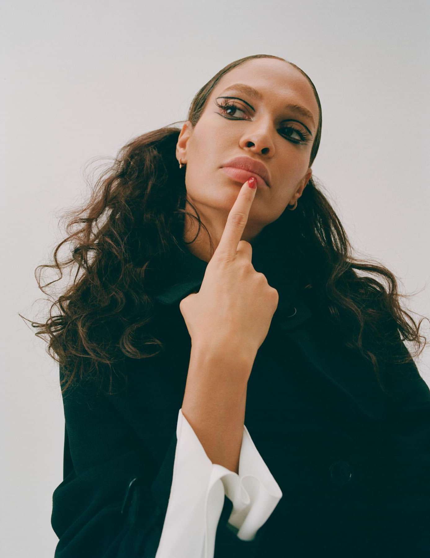 Portrait of an adult female with long wavy pigtails, wearing a dark jacket with white cuffs, posing with finger near her lips.