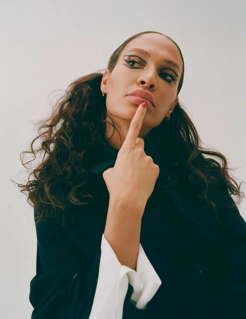 Portrait of an adult female with long wavy pigtails, wearing a dark jacket with white cuffs, posing with finger near her lips.