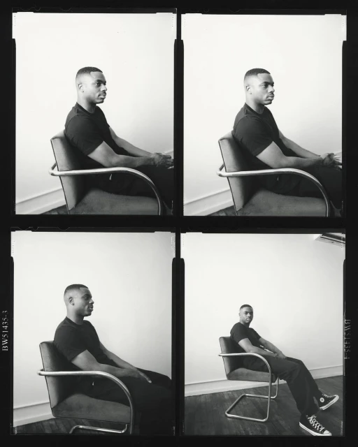 Black and white portrait of a man in a black shirt sitting in a chair with metal armrests against a bright white background.