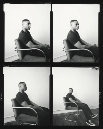 Black and white portrait of a man in a black shirt sitting in a chair with metal armrests against a bright white background.