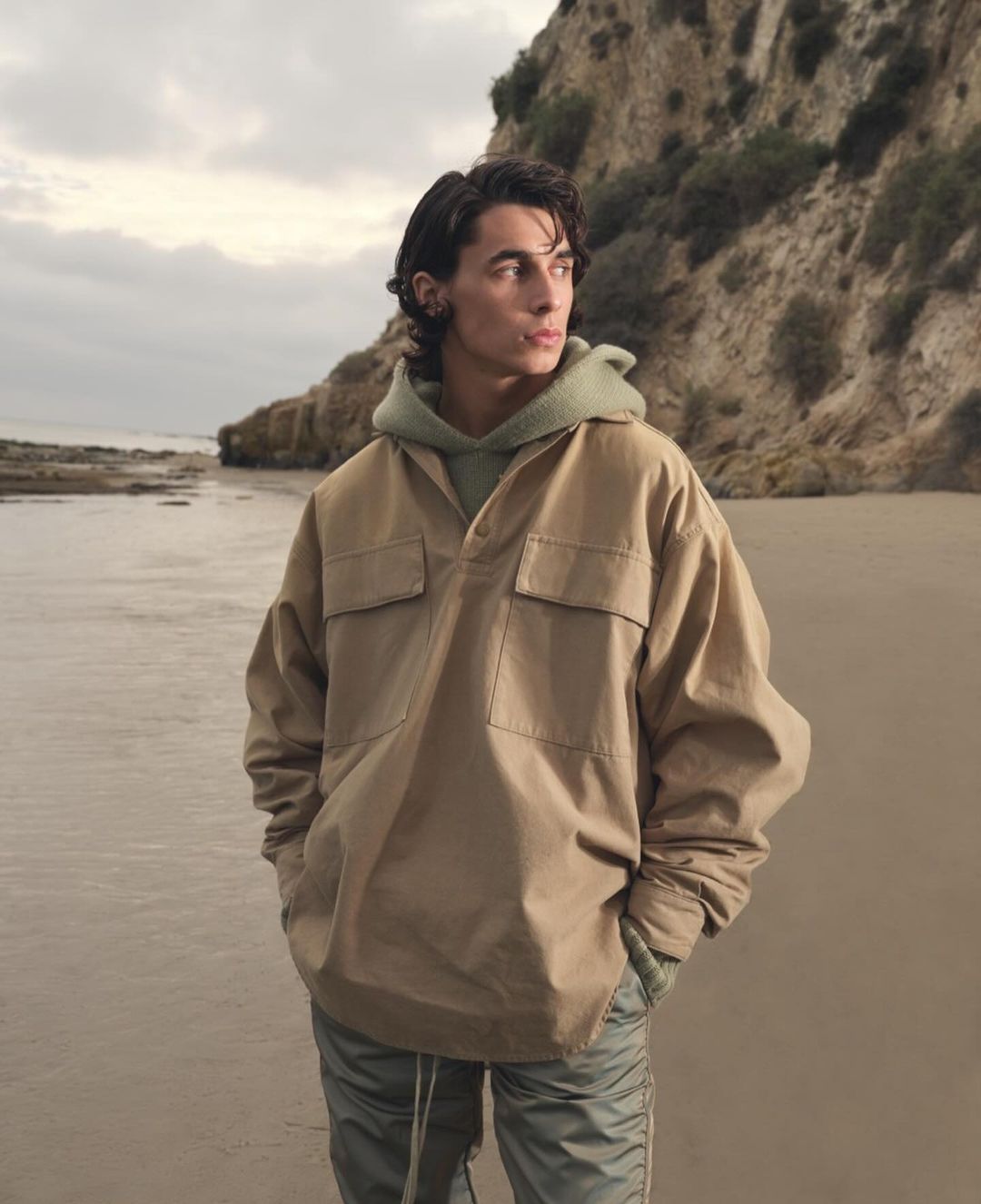 Man in a tan jacket and green hoodie standing on a sandy beach with a rocky cliff in the background looking off into the distance.