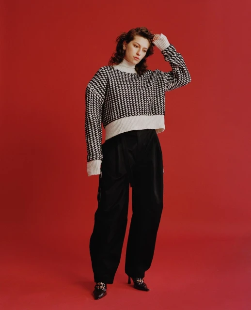Young woman poses in studio wearing black and white patterned sweater with turtleneck, black pants, and black high-heeled shoes on red backdrop.