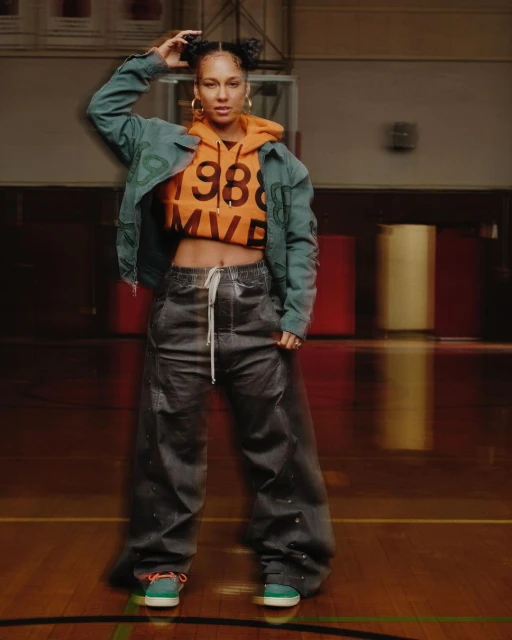 Singer Alicia Keys poses indoors wearing a bright orange hoodie with the text '1988 MVP' and loose dark grey pants and teal sneakers.