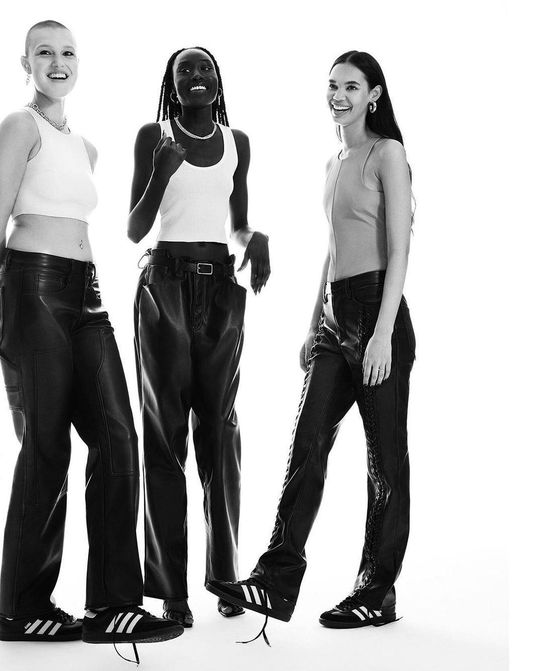 Three smiling women in white tops and black leather pants and Adidas sneakers pose against a white backdrop in a fashion shot.