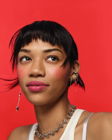 Close-up studio portrait of a young adult female with black hair, wearing jewelry and makeup, gazing upward against a solid red backdrop.