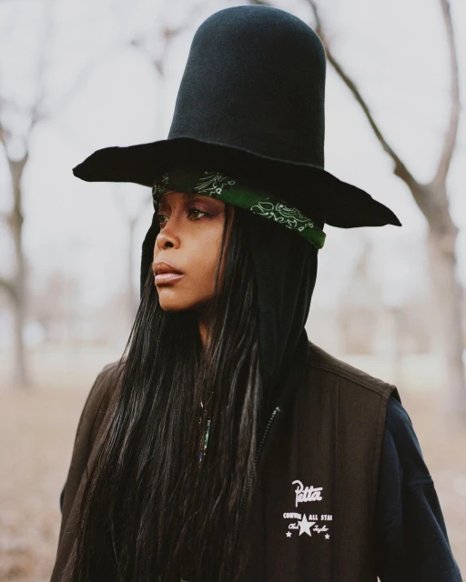 Woman with dark hair wearing a large black hat and bandana stands outdoors in a brown vest over a hooded sweatshirt and neutral environment.