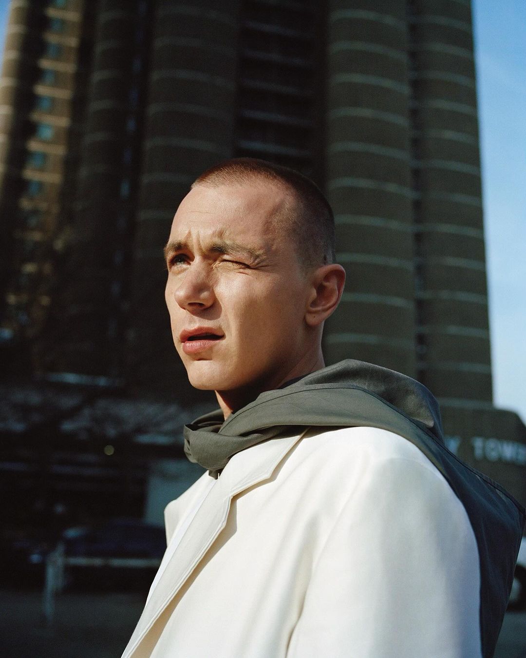 Portrait of a young adult man with a short buzzcut wearing a white coat, standing in an urban outdoor setting with a tall building backdrop.