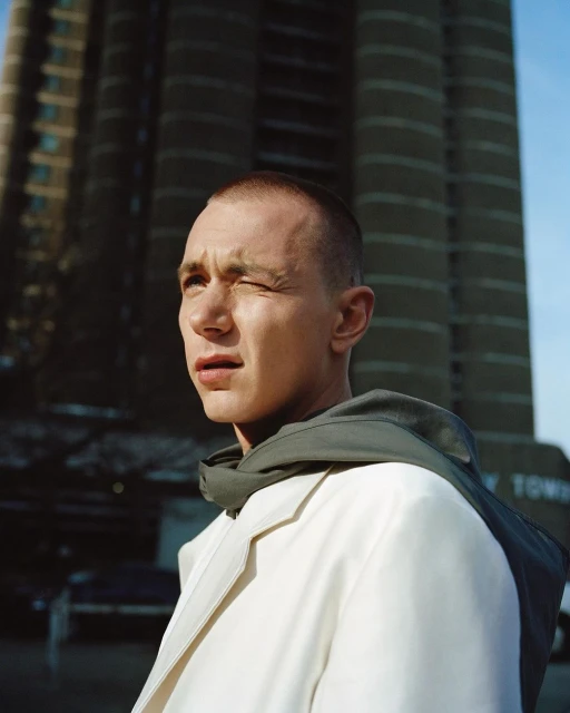 Portrait of a young adult man with a short buzzcut wearing a white coat, standing in an urban outdoor setting with a tall building backdrop.