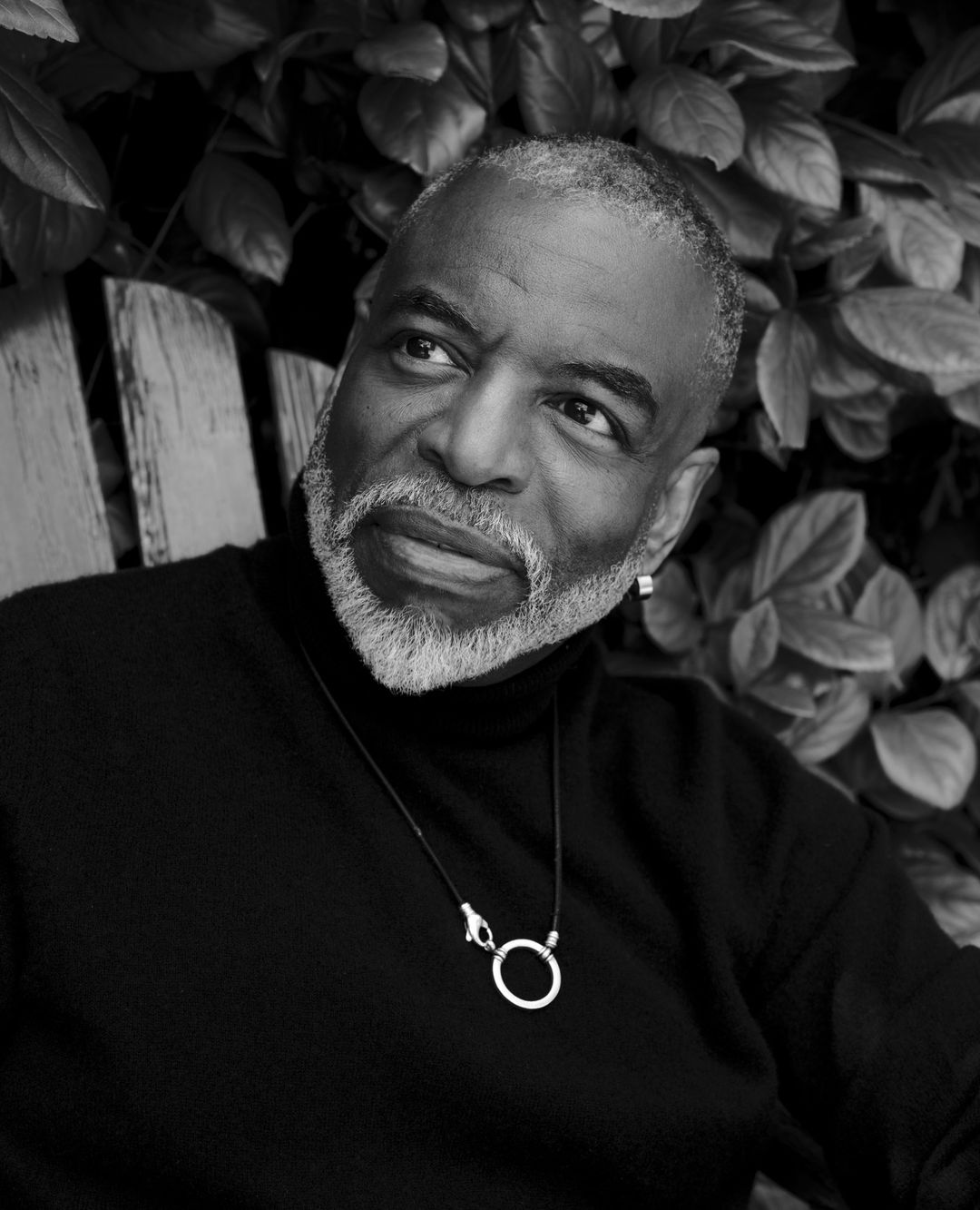 Portrait of an African American man in black turtleneck sweater. He's wearing a silver pendant necklace and has grey facial hair. Outdoor setting.