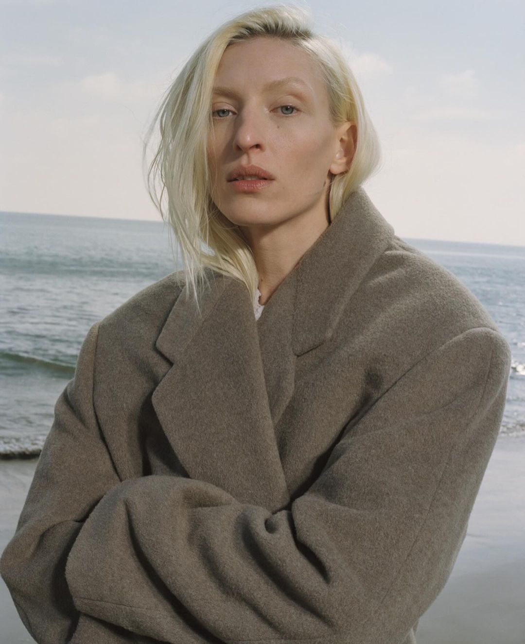 Fashion portrait of a blonde woman in a gray coat, posing outdoors near the ocean, showcasing neutral tones and a serene coastal backdrop.