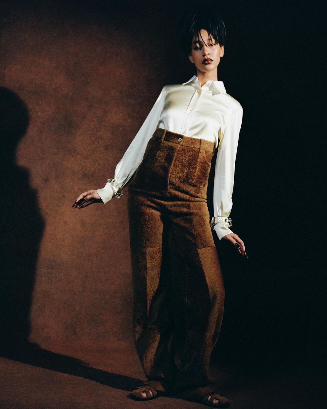 Adult female poses in a studio wearing a white satin blouse and brown textured wide-leg pants, against a brown background with soft shadows.