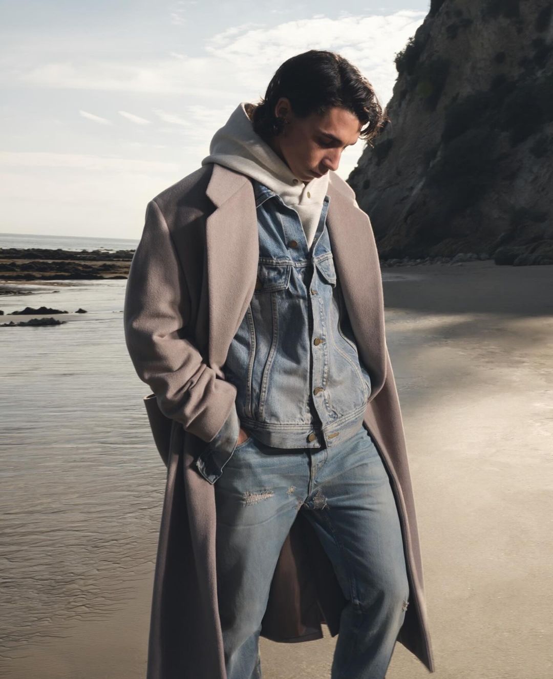 Young man in a stylish coat and denim jacket stands on a sandy beach next to the ocean, with cliffs in the background on a sunny day.
