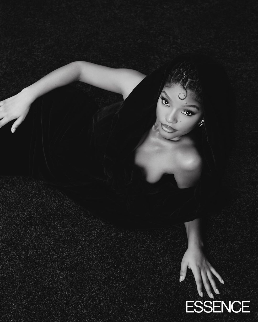 Black and white portrait of a woman with braided hair lying on a textured surface, wearing an off-the-shoulder dress, looking at the camera.