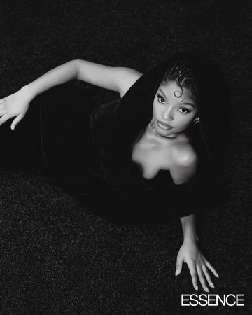 Black and white portrait of a woman with braided hair lying on a textured surface, wearing an off-the-shoulder dress, looking at the camera.