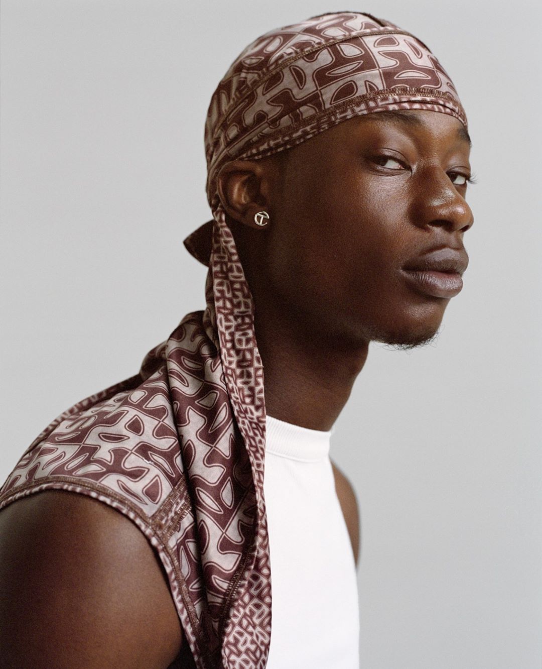 Portrait of a stylish African-American man wearing a patterned durag and tank top against a plain grey backdrop in a studio shot.